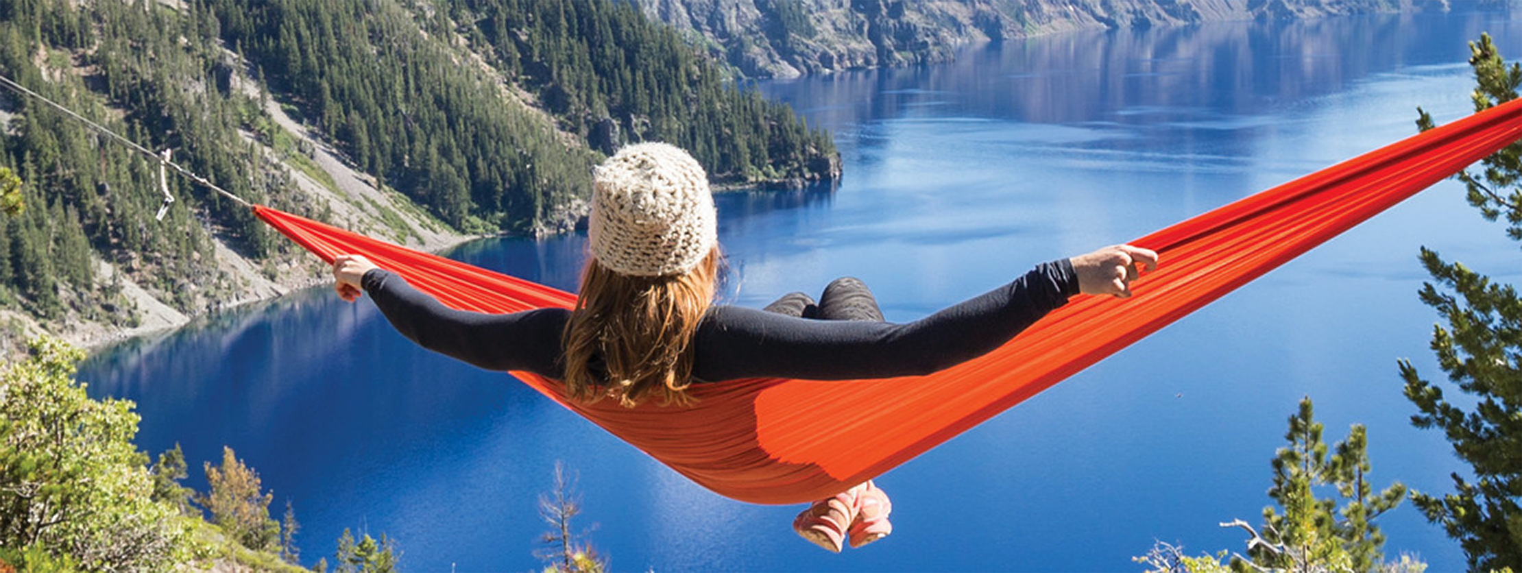 Person relaxing in an orange hammock suspended over a scenic lake surrounded by forested cliffs, wearing a knit beanie and facing the water