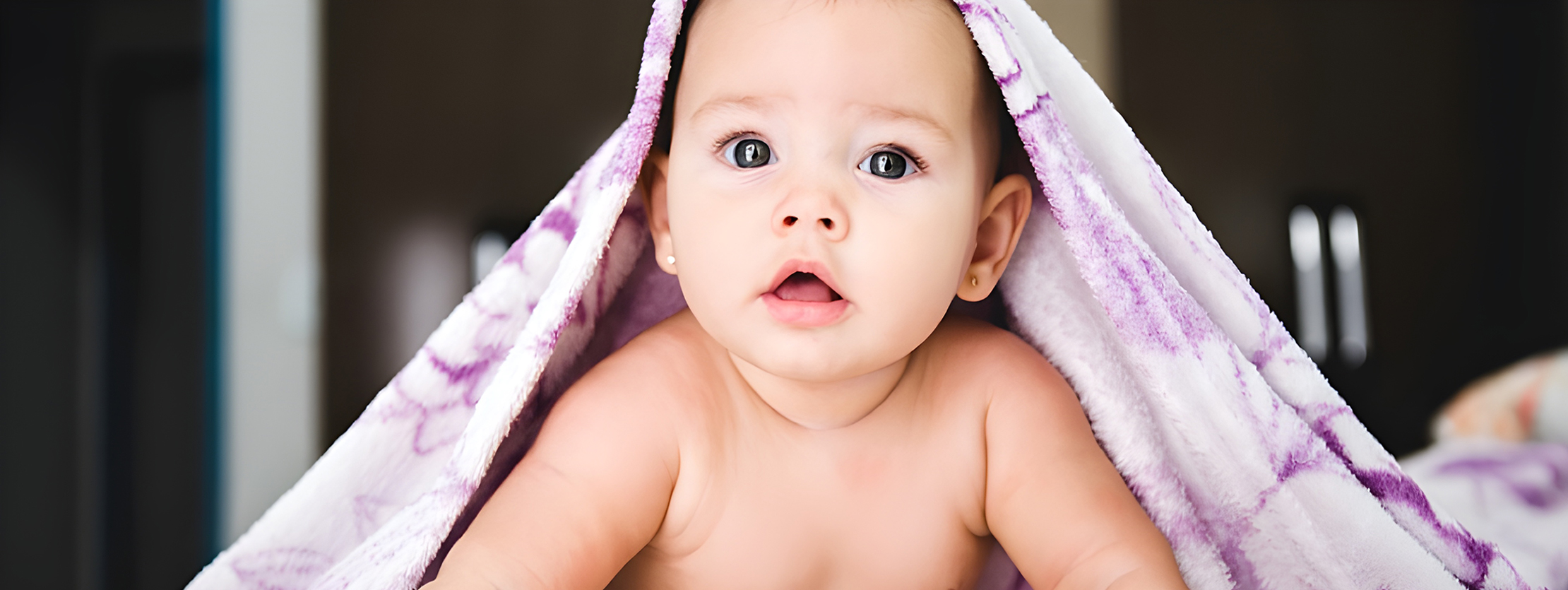 Baby with bright eyes, wrapped in a soft, purple-patterned blanket, looking curiously at the camera