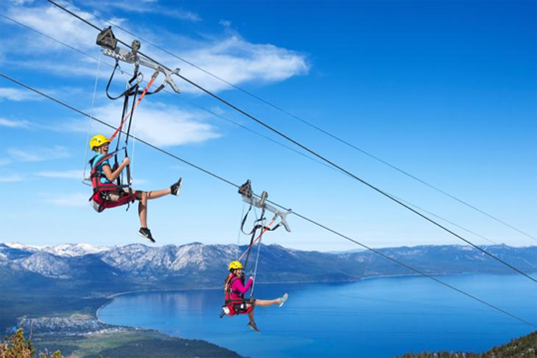 Two people ziplining high above a large blue lake with mountain ranges in the background, wearing helmets and harnesses under a clear blue sky.