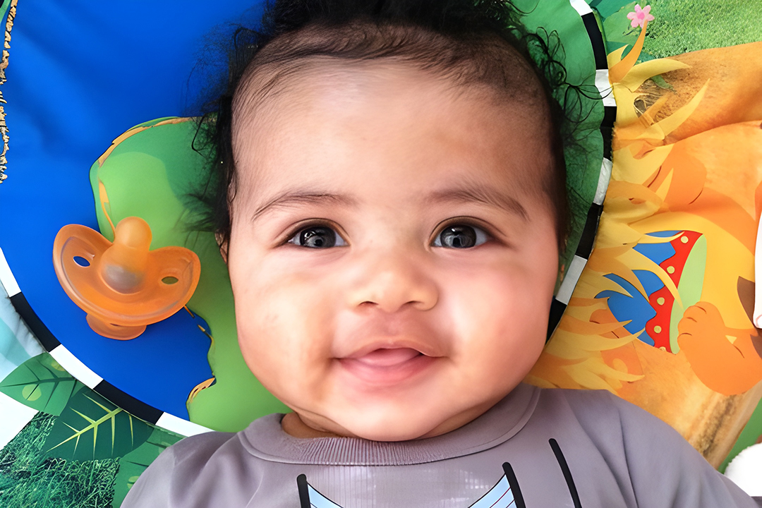 Happy baby lying on a colorful playmat with a bright orange pacifier nearby, smiling up at the camera