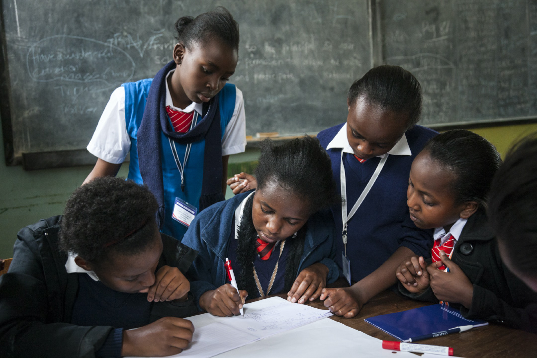 Group of young students in school uniforms collaborating over a notebook in a classroom with a chalkboard in the background.
