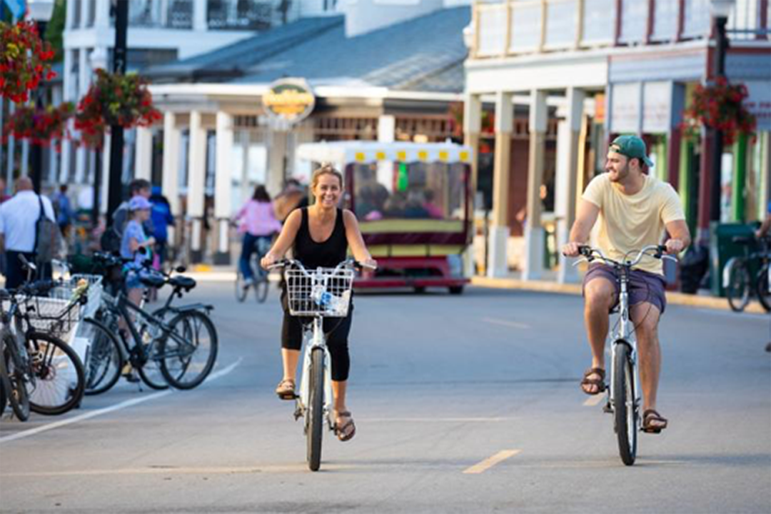 Two people riding bikes and smiling on a charming town street lined with shops, pedestrians, and parked bicycles on a sunny day.