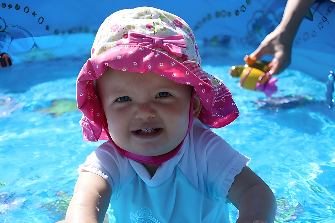 Cheerful baby in a pink floral sun hat and white rash guard, enjoying a splash in a bright blue kiddie pool